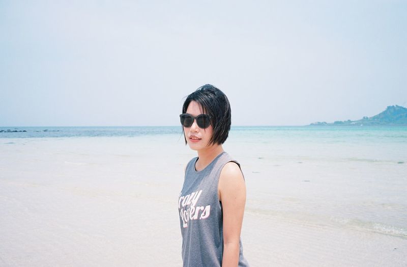 Young woman wearing sunglasses standing at beach against clear sky