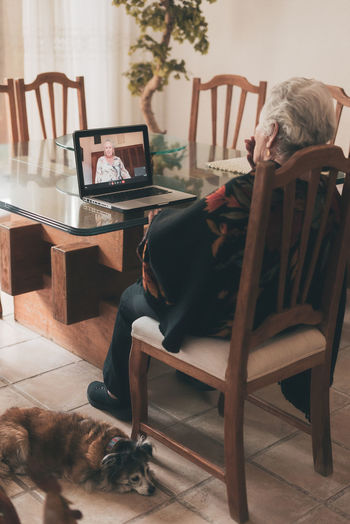 Back view of unrecognizable elderly female with short gray hair sitting on chair and listening goodbye of sister while making video call via netbook at home