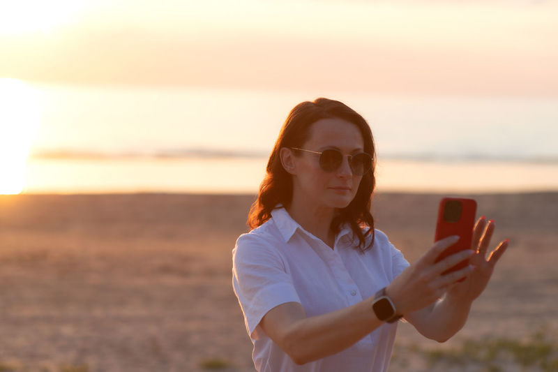Young woman using mobile phone while standing against sky during sunset