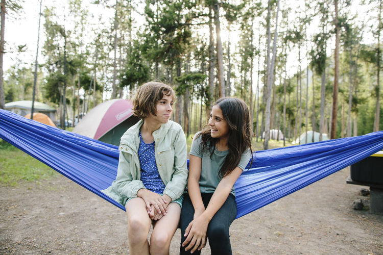 Sisters talking while sitting on hammock against trees in forest
