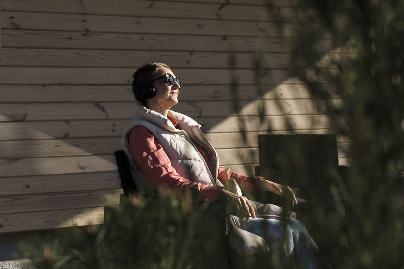 Woman wearing sunglasses and headphones sitting on veranda in sunshine