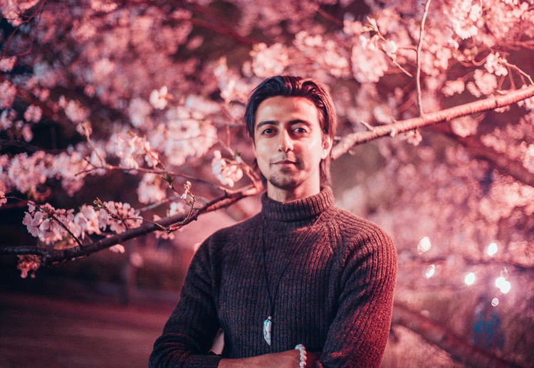 Portrait of confident young man with arms crossed standing by cherry tree at night