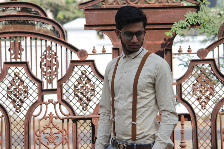 Thoughtful young man wearing suspenders standing against gate
