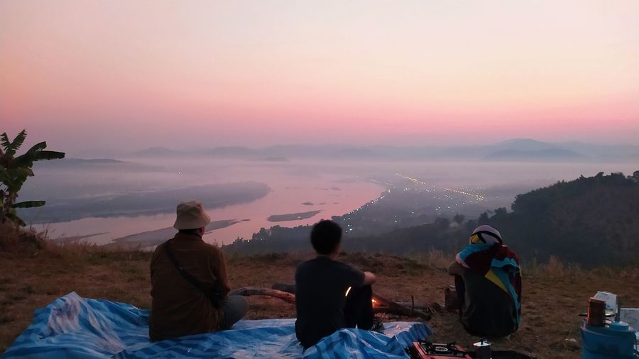 Rear view of people sitting on mountain against sky during sunset