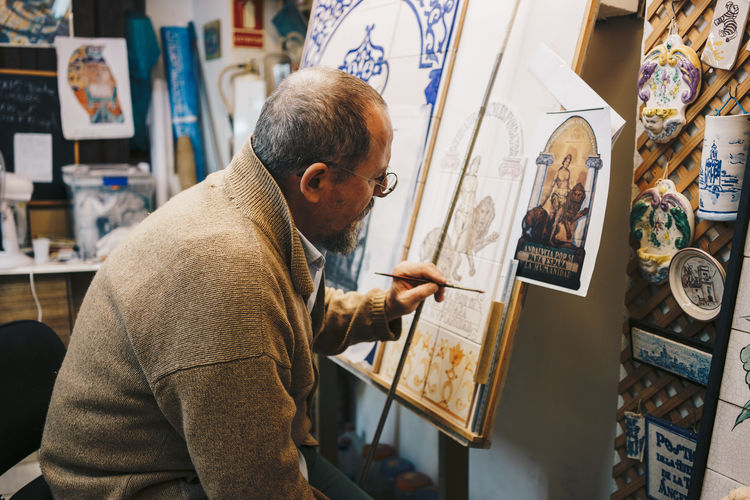 From above side view of aged focused artist in casual clothes sitting in front of paint easel and drawing patterns on ceramic tile in workshop among ceramic items