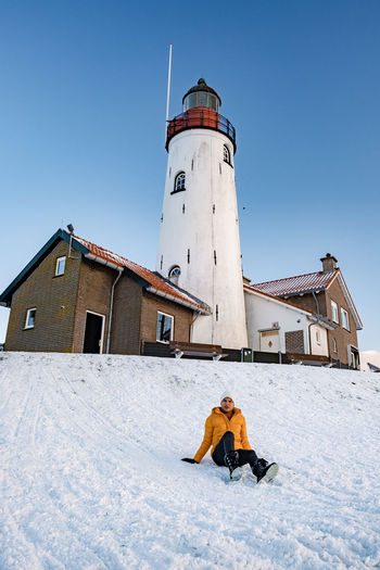 Full length of couple sitting on snow covered land against lighthouse