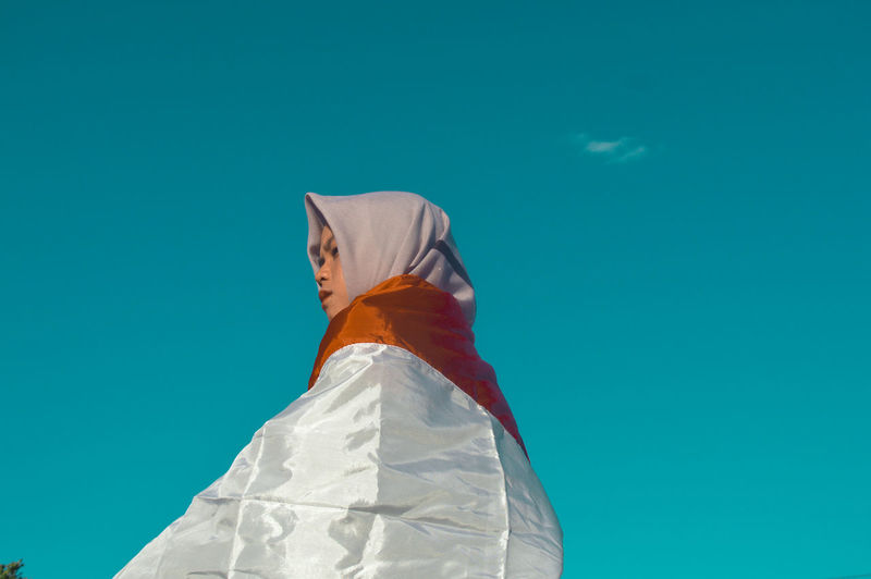 Low angle view of woman with indonesia flag standing against clear blue sky