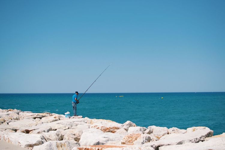 Rear view of man fishing by sea against clear blue sky