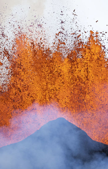 Splashes of hot orange lava erupting from volcanic mountain peak surrounded with smoke in iceland
