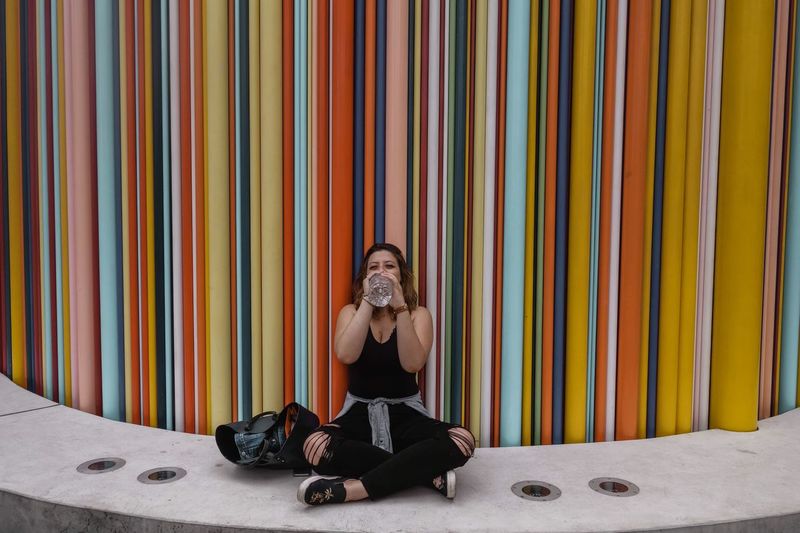 Full length of woman drinking water while sitting against multi colored wall