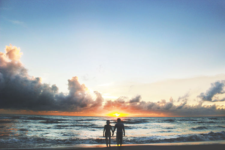 Rear view of couple holding hands while standing at beach against cloudy sky during sunset