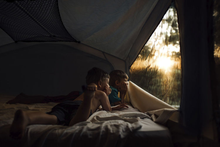 Thoughtful brothers looking through window while lying in tent