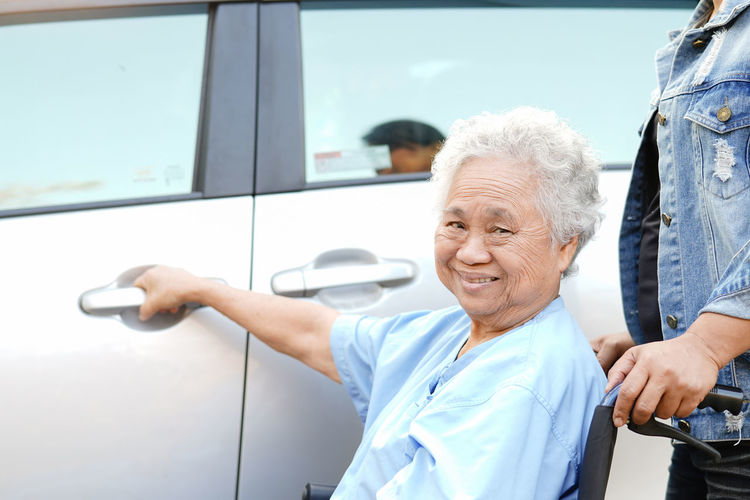 Portrait of smiling senior female patient sitting on wheelchair opening car door