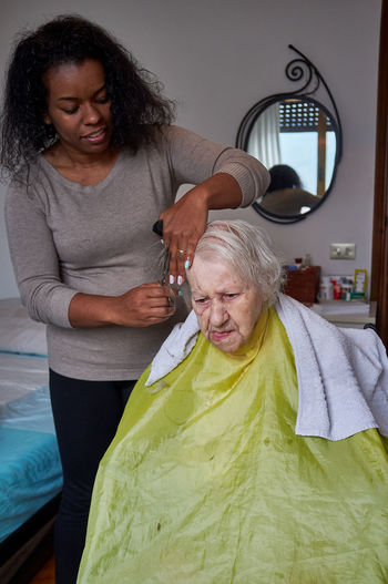 Caring african american woman caregiver, cutting her elderly woman hair at home