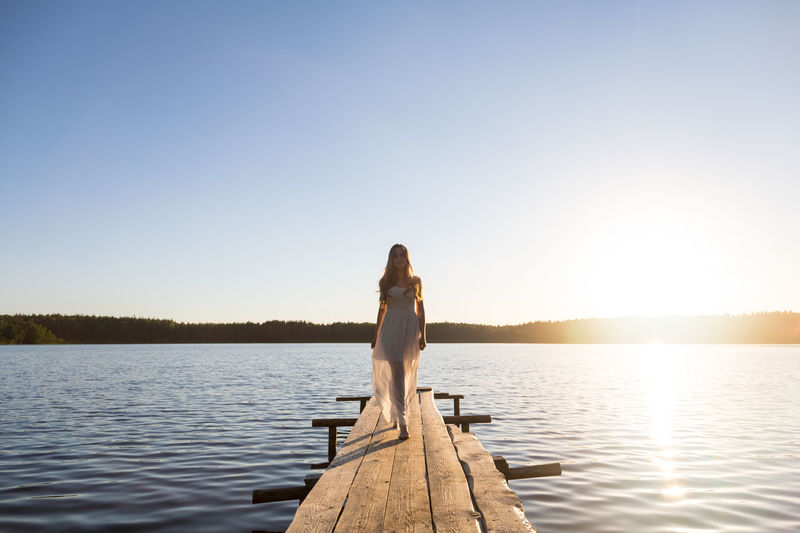 Rear view of woman standing by lake against clear sky during sunset