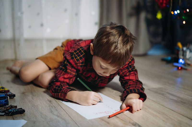 Portrait of preschooler boy in plaid shirt laying on the floor writing christmas letter to santa