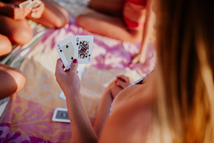High angle back view of unrecognizable young woman holding 2 and queen of spades cards while playing game with friends on beach