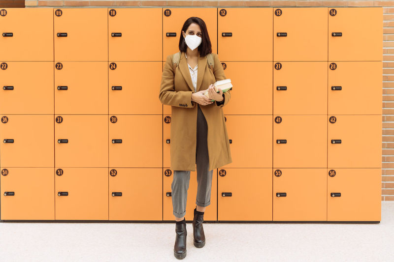Female student wearing protective mask standing with pile of books near lockers in university corridor and looking at camera during coronavirus epidemic