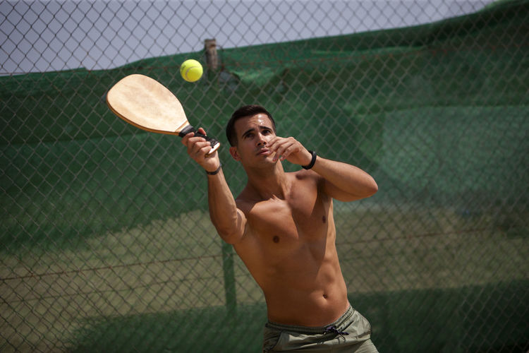 Shirtless young man playing tennis while standing against chainlink fence