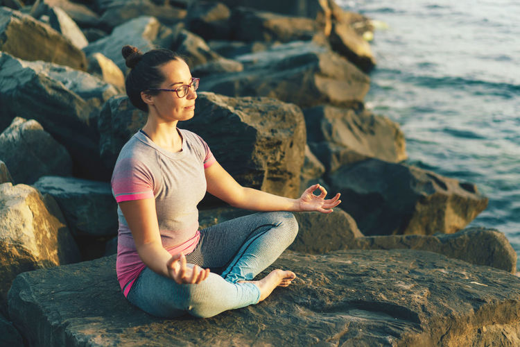 Full length of woman meditating while sitting on rock at sea