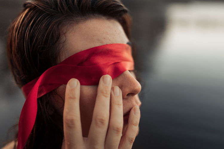 Side view of anonymous gentle female tourist in red blindfold touching face against water in daylight