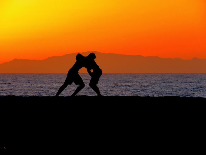 Silhouette men wrestling on shore at beach against orange sky during sunset
