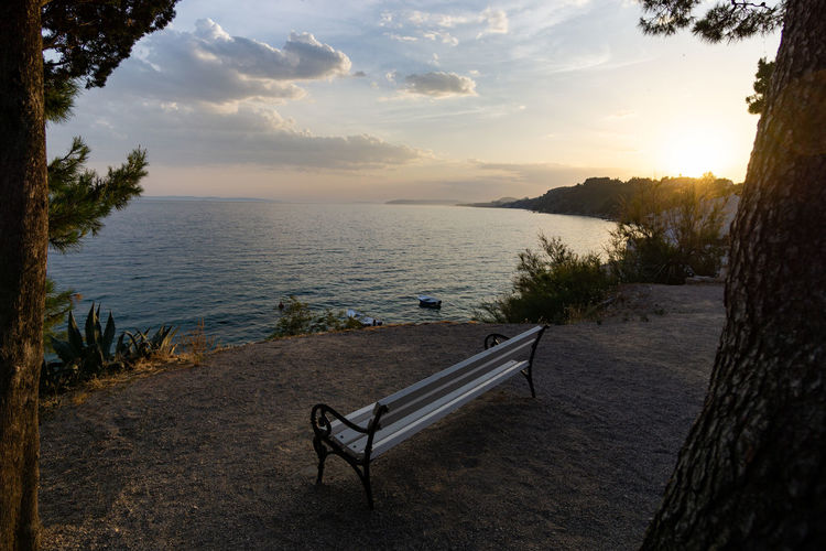 Empty bench by sea against sky during sunset