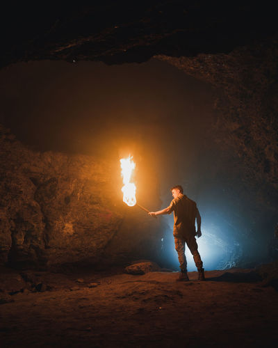 Back view of young male speleologist with flaming torch standing in dark narrow rocky cave while exploring subterranean environment