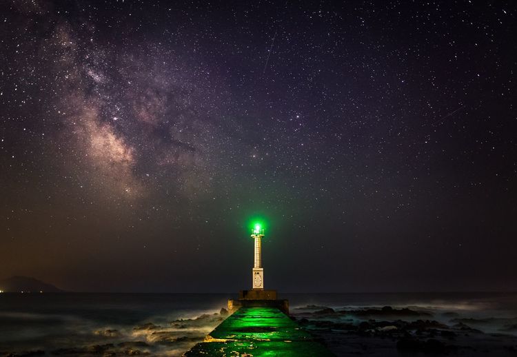 Pier leading towards illuminated lighthouse in front of sea at night