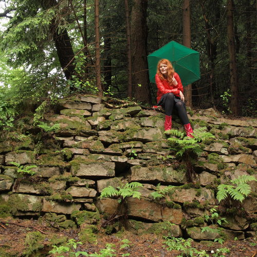 Woman in raincoat with umbrella sitting on stone wall at forest