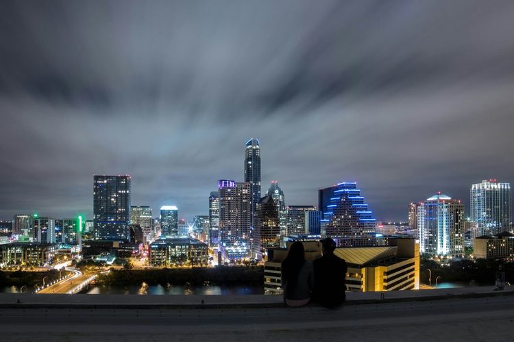 Man and woman looking at illuminated cityscape while sitting on terrace at night