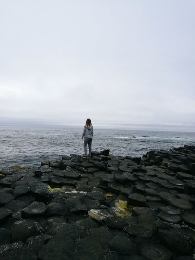 Rear view of woman standing on basalt columns by sea against sky at giant causeway