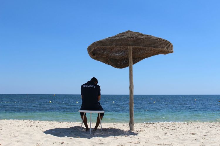 Rear view of lifeguard sitting under parasol at beach against sky on sunny day