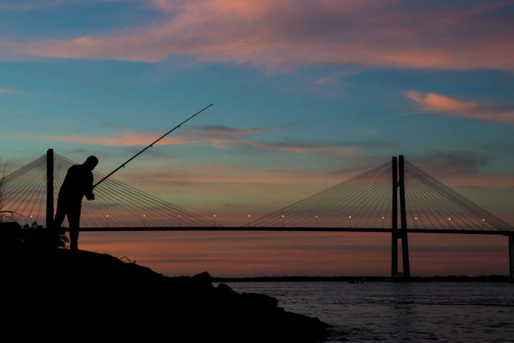 Silhouette man fishing in river against rosario-victoria bridge during sunset