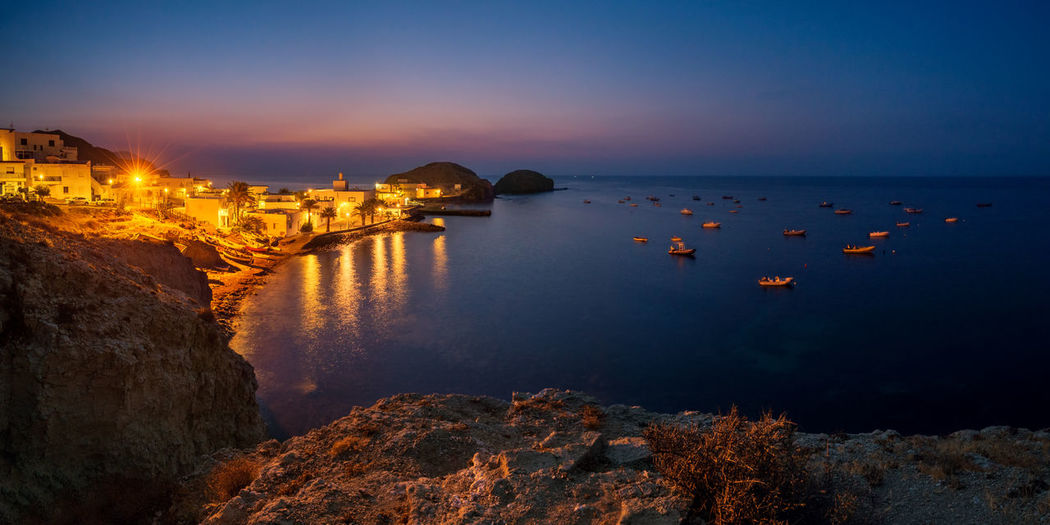 Scenic view of sea by illuminated city against sky at sunset