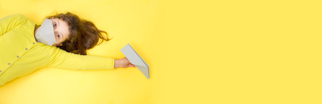 High angle portrait of girl wearing mask lying down on yellow background