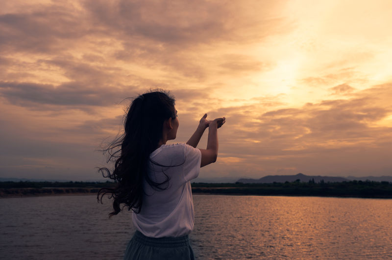 Rear view of woman praying while standing by river during sunset