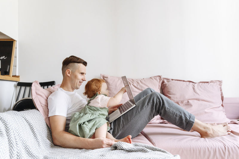 Man with baby daughter working on laptop on sofa
