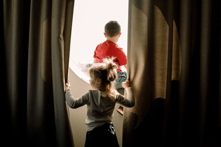 Rear view of girl holding curtain while brother sitting on window in hotel room