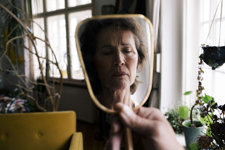 Reflection of upset senior woman on hand mirror at home