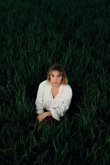 Peaceful young female in retro styled white blouse sitting amidst tall green grass and looking at camera while resting in summer evening in countryside