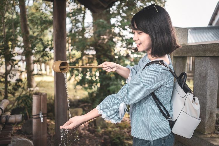 Side view of smiling woman pouring water from ladle at shrine