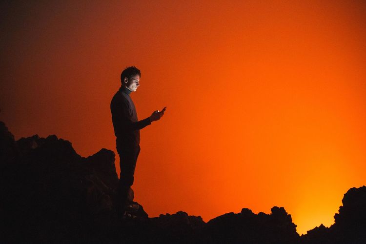 Side view of man using mobile phone while standing on silhouette rocks during sunset