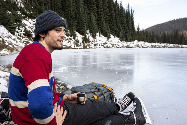 Young hockey player resting near frozen lake and drinking hot beverage