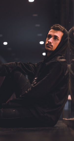 Side view of young man wearing hooded jacket sitting on floor in dark room