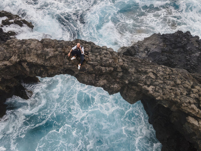 Mature man sitting on rock arch above sea