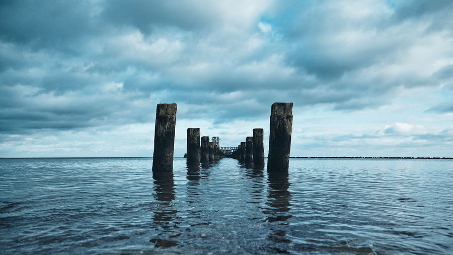Wooden posts in sea against cloudy sky