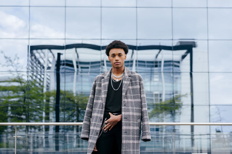 Confident young african american male teenager model in trendy tartan coat and stylish accessories looking at camera while standing against contemporary building with glass wall
