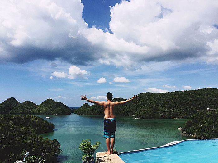 Rear view of shirtless with arms outstretched standing by infinity pool and sea against cloudy sky