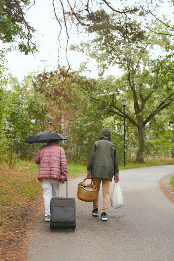 Rear view of grandmother and grandson with luggage walking on road in park during rainy season